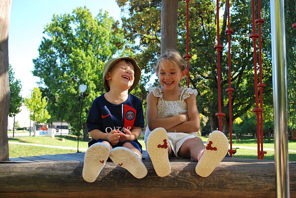Dve smejúce sa deti - chlapec a dievča - sedia na detskom ihrisku. Majú obuté topánky VIVOBAREFOOT.