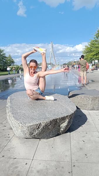 Je leto. Autorka článku je v jogovej pozícii v mestskom prostredí na veľkom kameni, sedí s jednou nohou zdvihnutou a natiahnutou k hlave. Na nohách má položené zariadenie VIVOBAREFOOT Motus Flex.